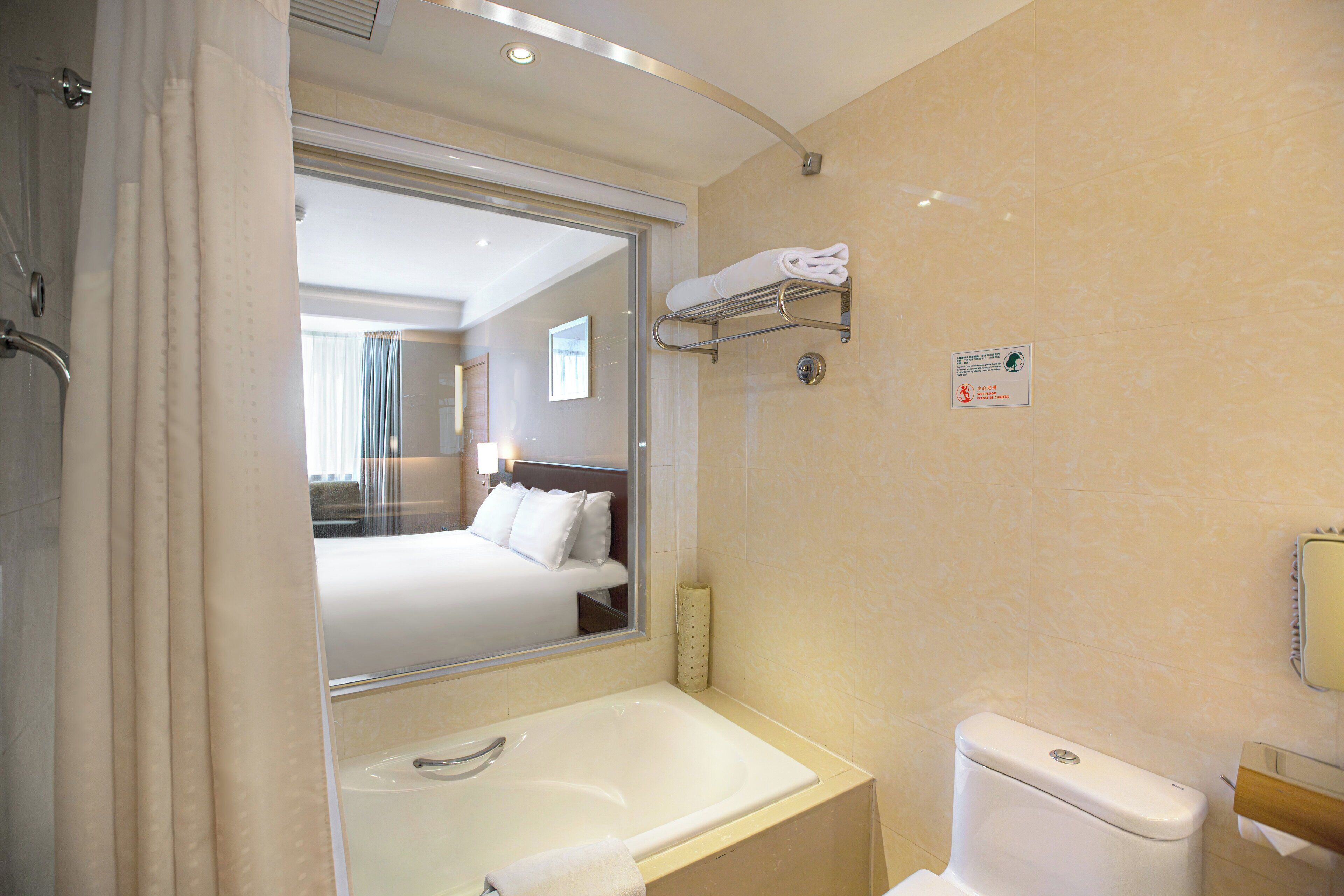 Een hotelbadkamer met een badkuip, toilet en een spiegel die een slaapkamer met een bed en raam weerkaatst.