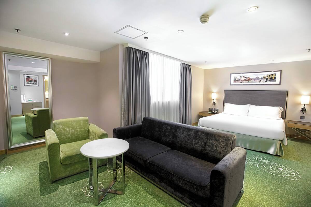 A hotel room with a bed, a green armchair, a black sofa, a small round table, and a mirror.