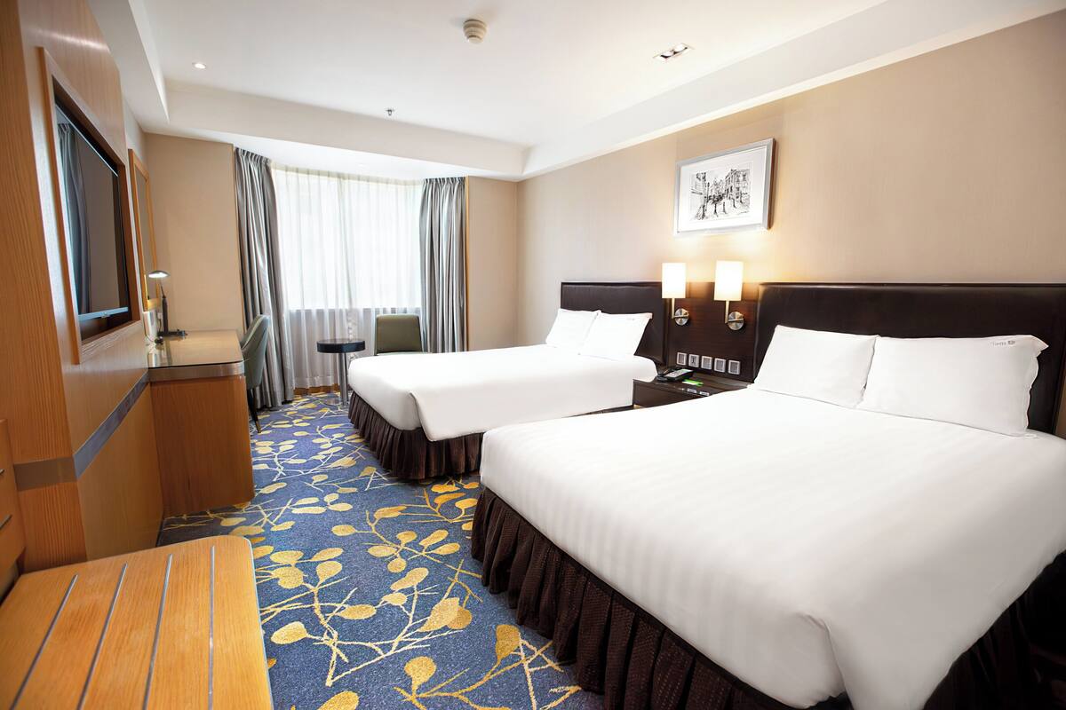 A hotel room with two beds, a desk, a TV, and a window with curtains.