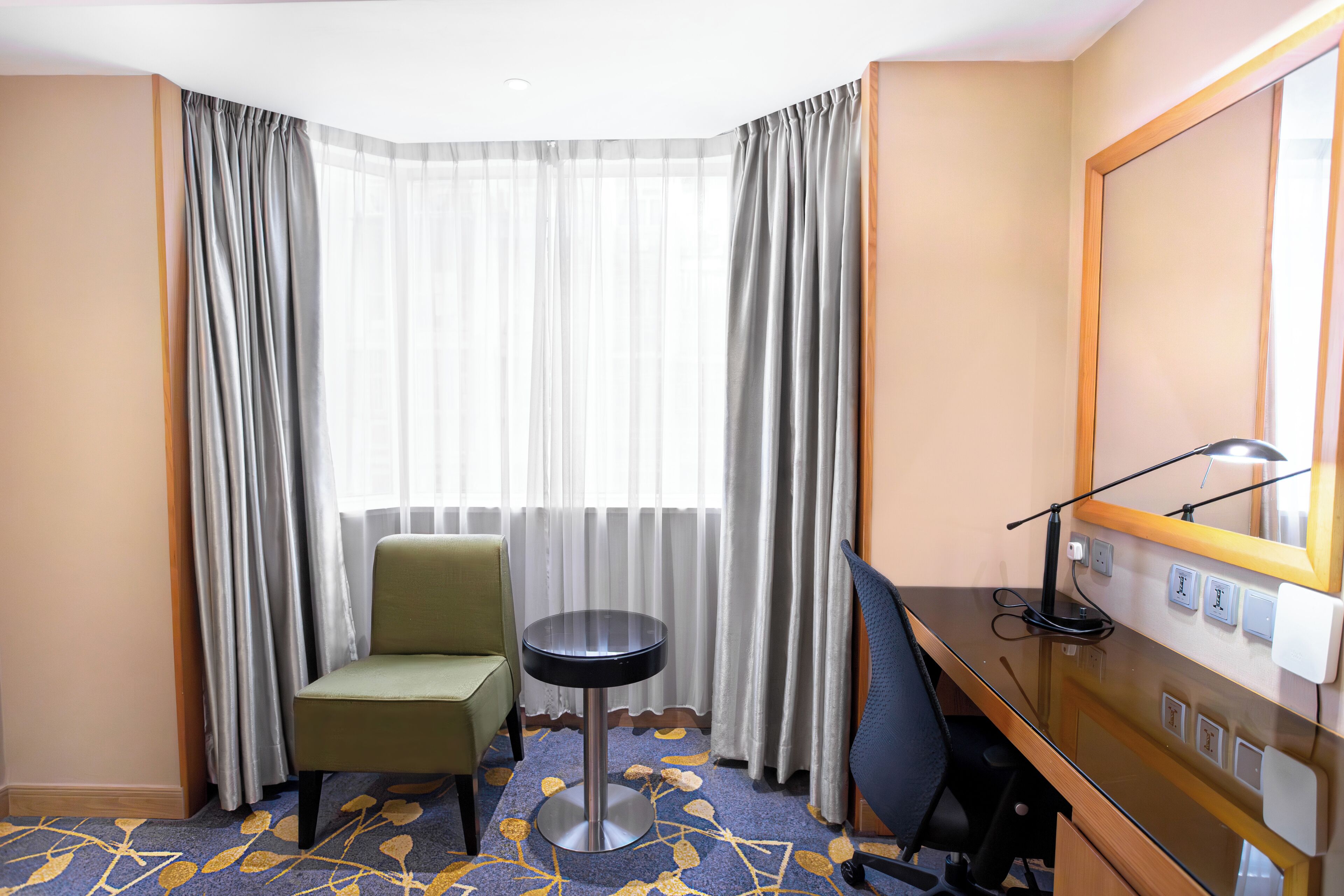 Een hotelkamer met een hoekbureau, een groene fauteuil, een klein rond tafeltje en een grote spiegel.