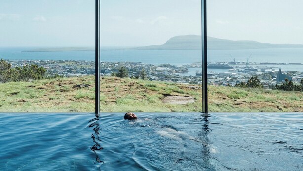 A person in a swimming pool with a view of a coastal cityscape and a distant mountain. - Hotel Føroyar (Tórshavn)