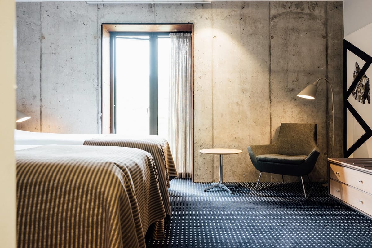 Une chambre d’hôtel avec deux lits, une petite table ronde, une chaise, un bureau avec des tiroirs et une grande fenêtre avec des rideaux.