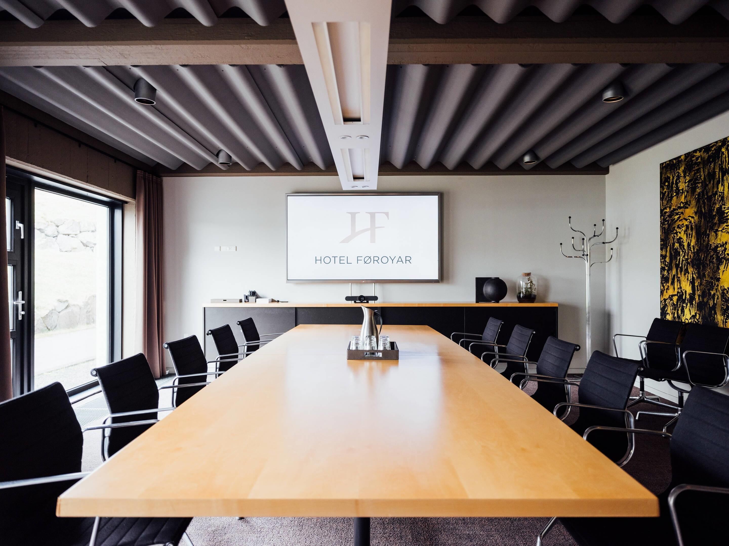 Una sala conferenze con un lungo tavolo, sedie, uno schermo per proiettore che mostra 'HOTEL FØROYAR' e un design moderno del soffitto.