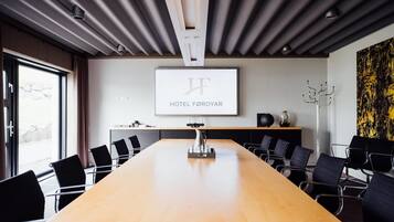 Una sala conferenze con un lungo tavolo, sedie, uno schermo per proiettore che mostra 'HOTEL FØROYAR' e un design moderno del soffitto.