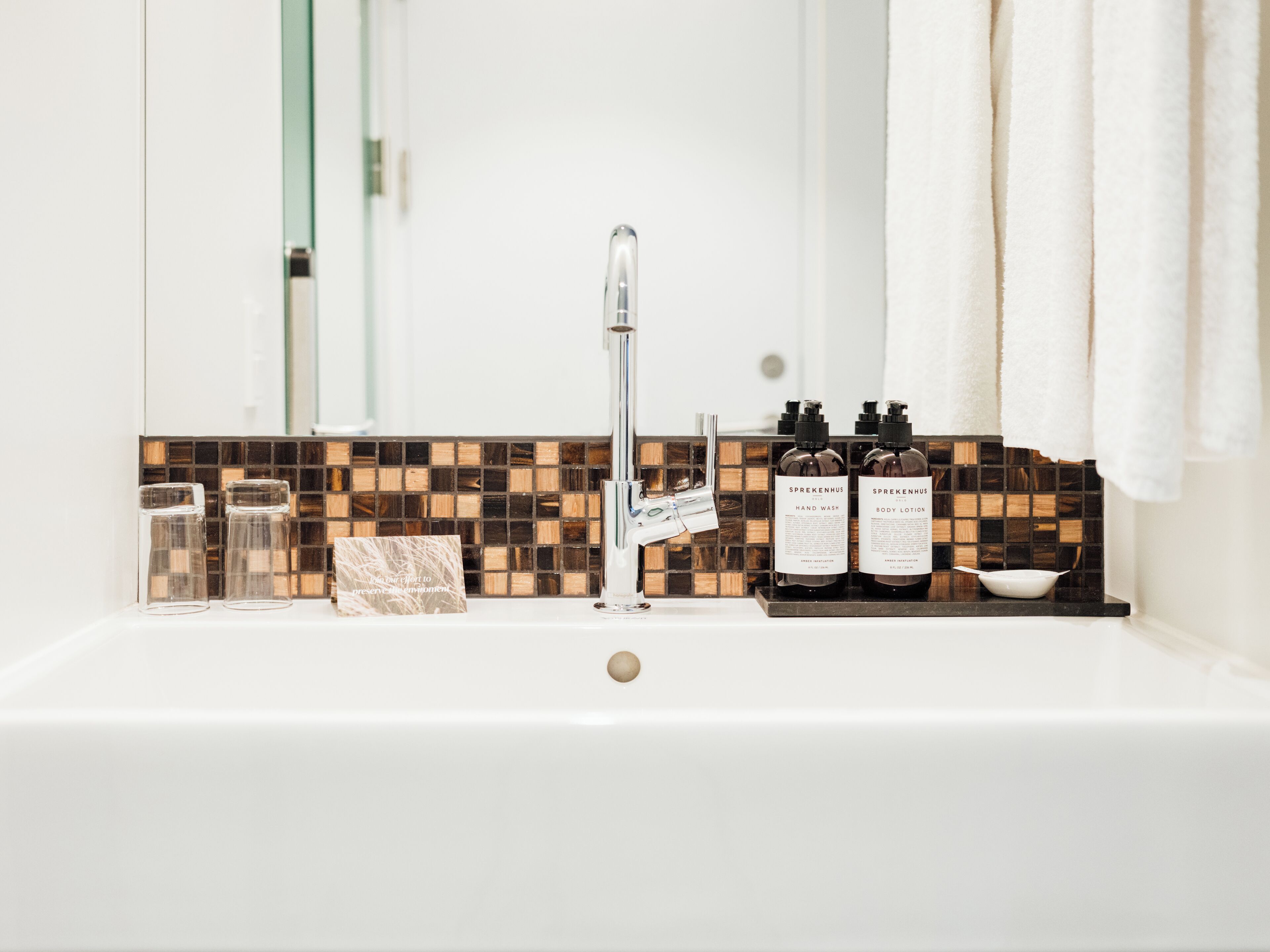 A bathroom with a white sink, a mirror, and a tiled backsplash. There are two bottles of hand wash and body lotion on the sink.