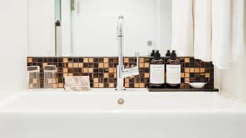 A bathroom with a white sink, a mirror, and a tiled backsplash. There are two bottles of hand wash and body lotion on the sink.