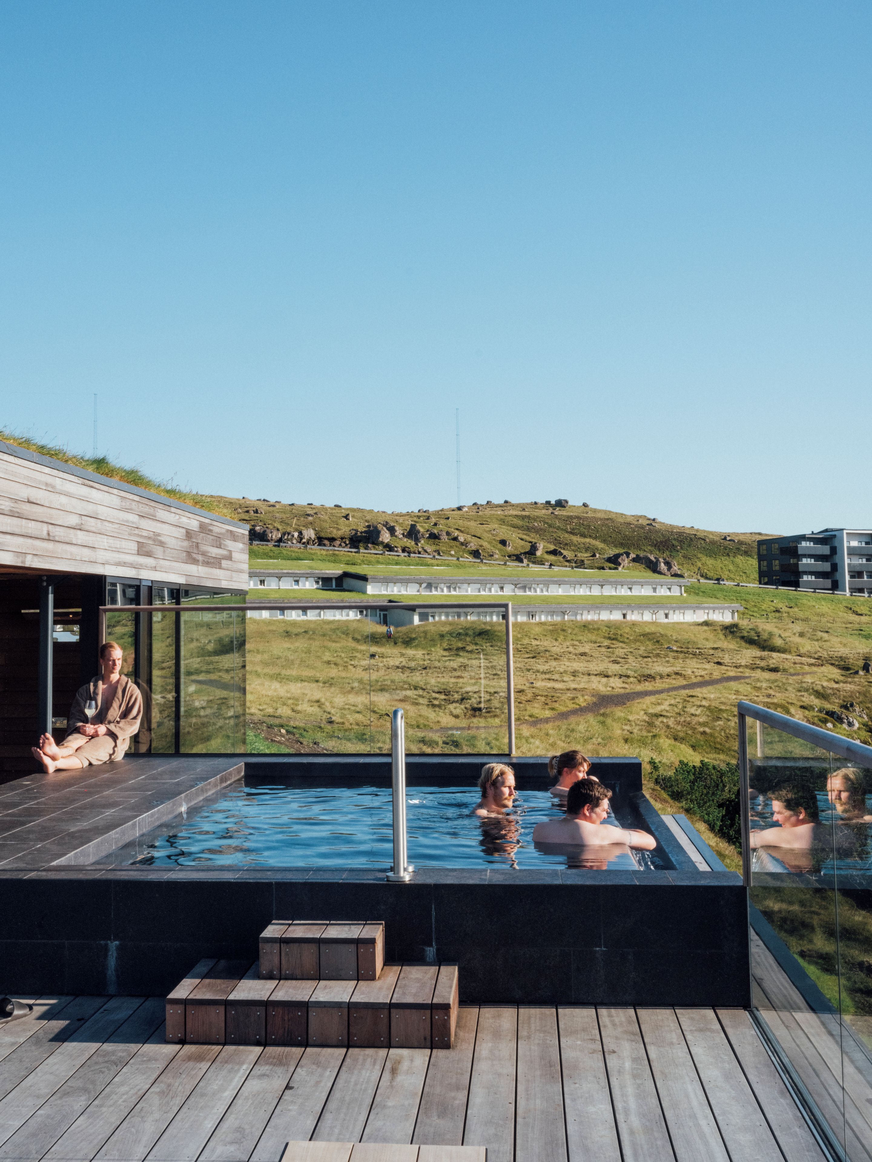 A modern outdoor swimming pool with clear glass walls, wooden deck, and a view of a grassy landscape.