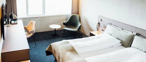A hotel room with a bed, a desk, a chair, a small table, and a window with curtains.