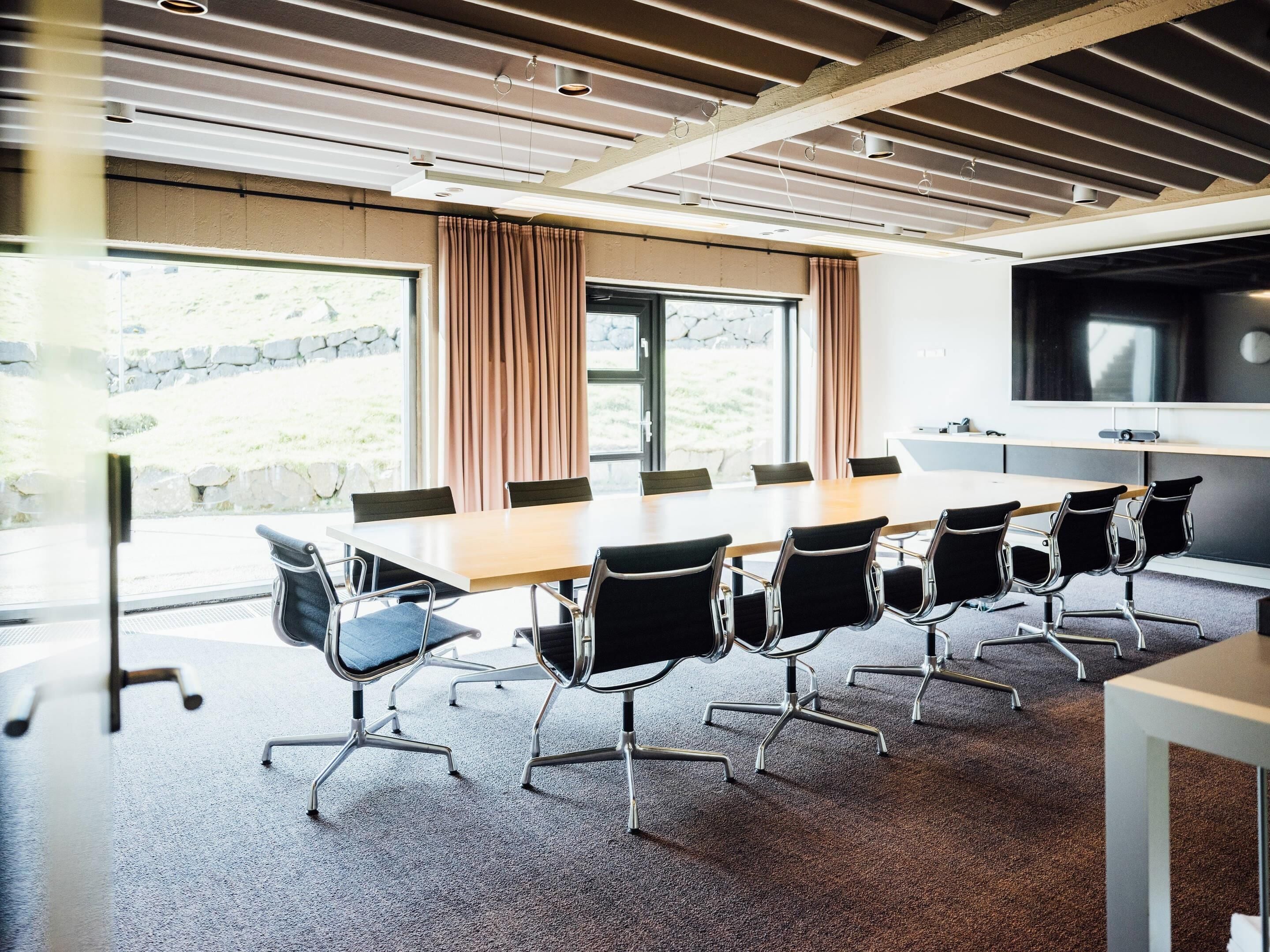 A conference room with a long table, black chairs, a large screen, and windows with curtains.