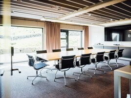 A conference room with a long table, black chairs, a large screen, and windows with curtains.