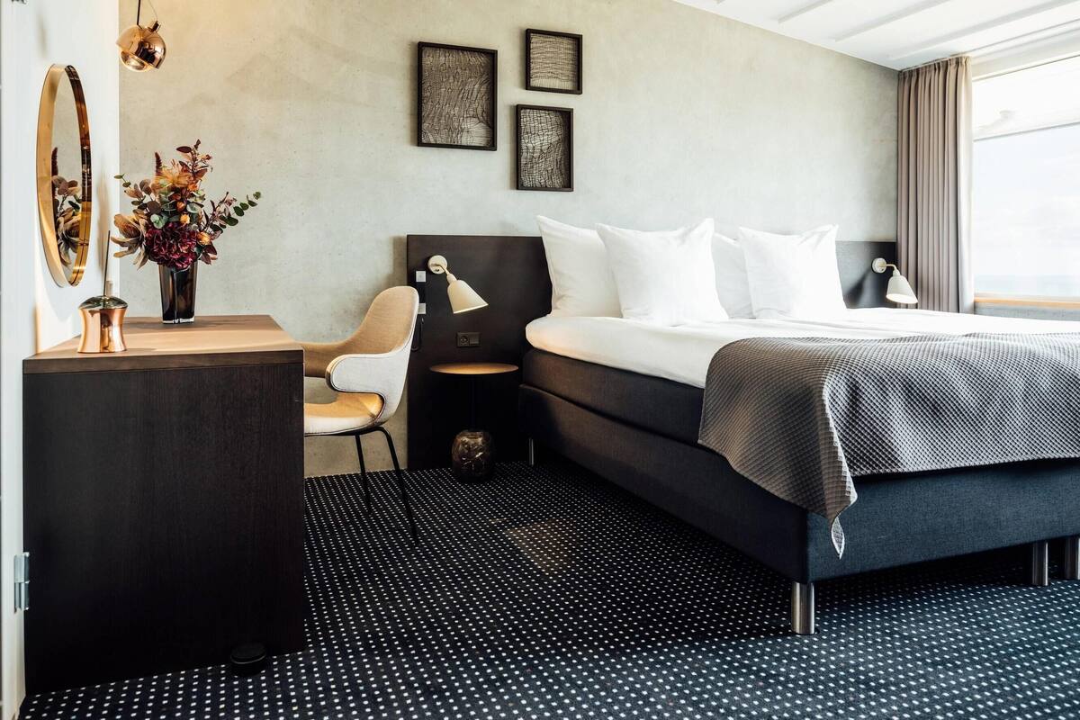 Une chambre d’hôtel moderne, équipée d’un lit, d’un bureau avec une lampe, d’une chaise et d’une petite table sur laquelle est posé un vase de fleurs.
