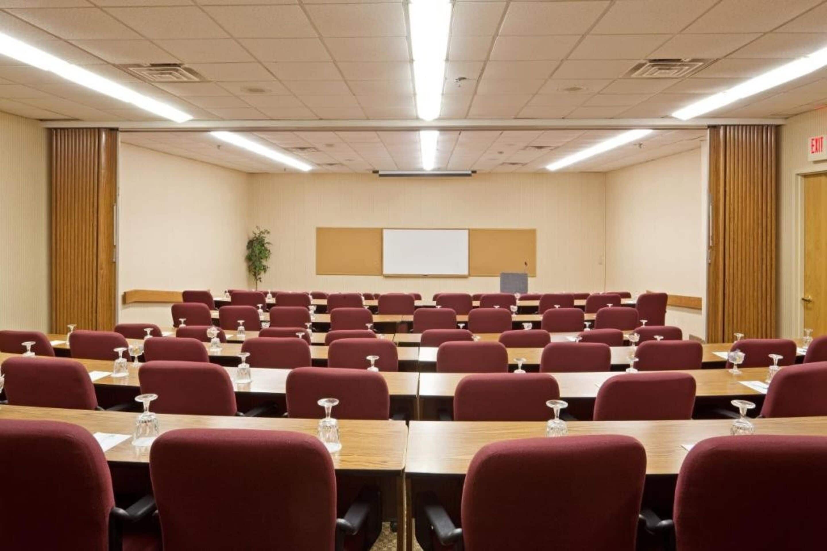 A conference room with rows of tables and chairs, a whiteboard, and an exit door.