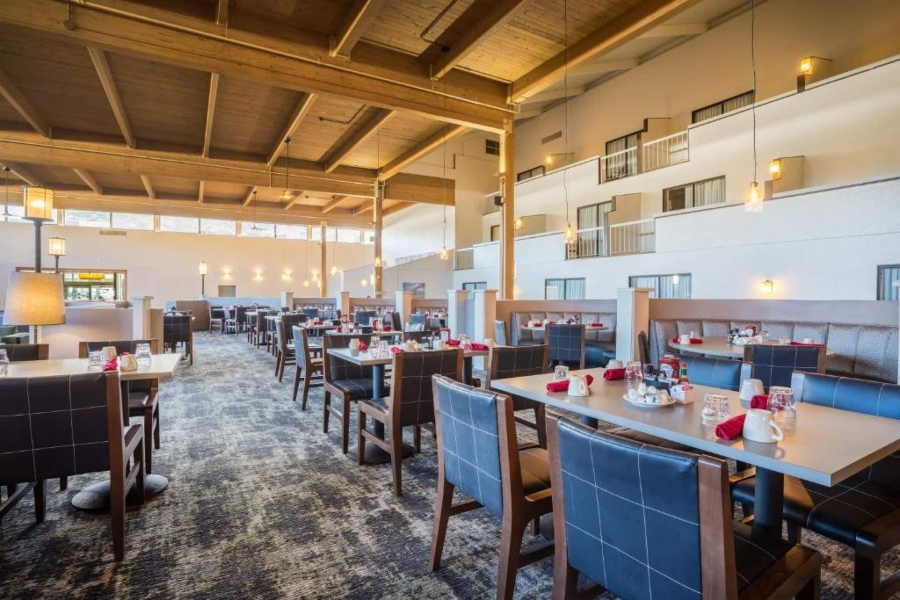 A spacious hotel dining area with multiple tables and chairs, high ceilings with exposed beams, and large windows allowing natural light.