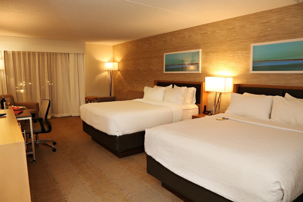 A hotel room with two beds, a desk, a chair, a lamp, and a painting on the wall.