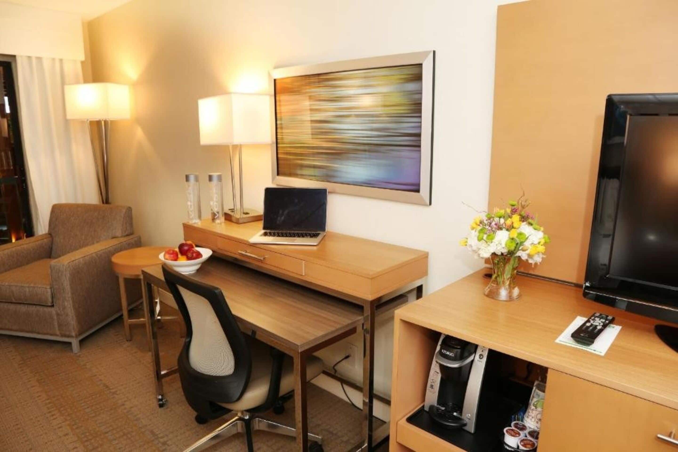 A hotel room with a desk, laptop, chair, TV, lamp, and a vase with flowers.