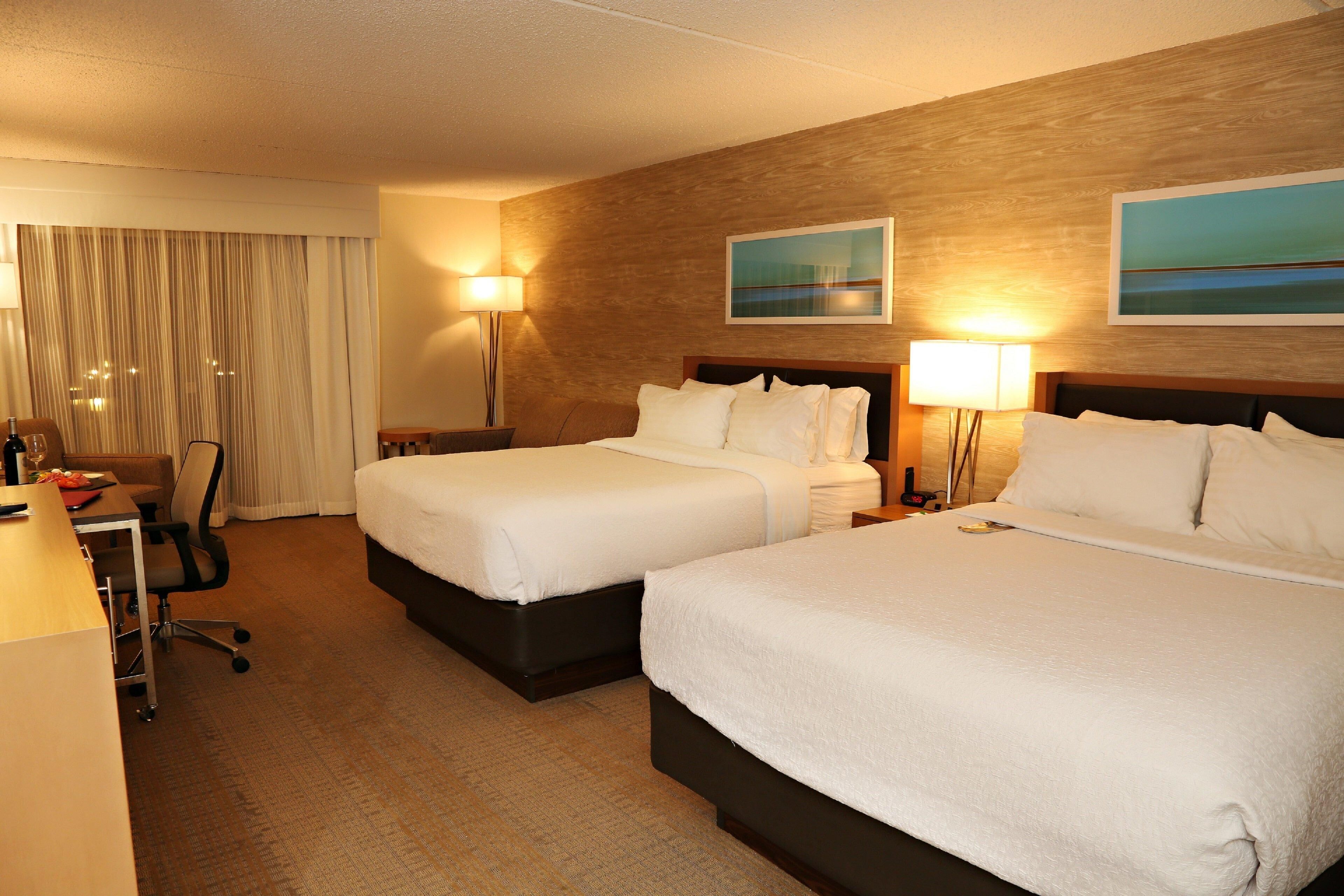 A hotel room with two beds, a desk, a chair, a lamp, and a painting on the wall.
