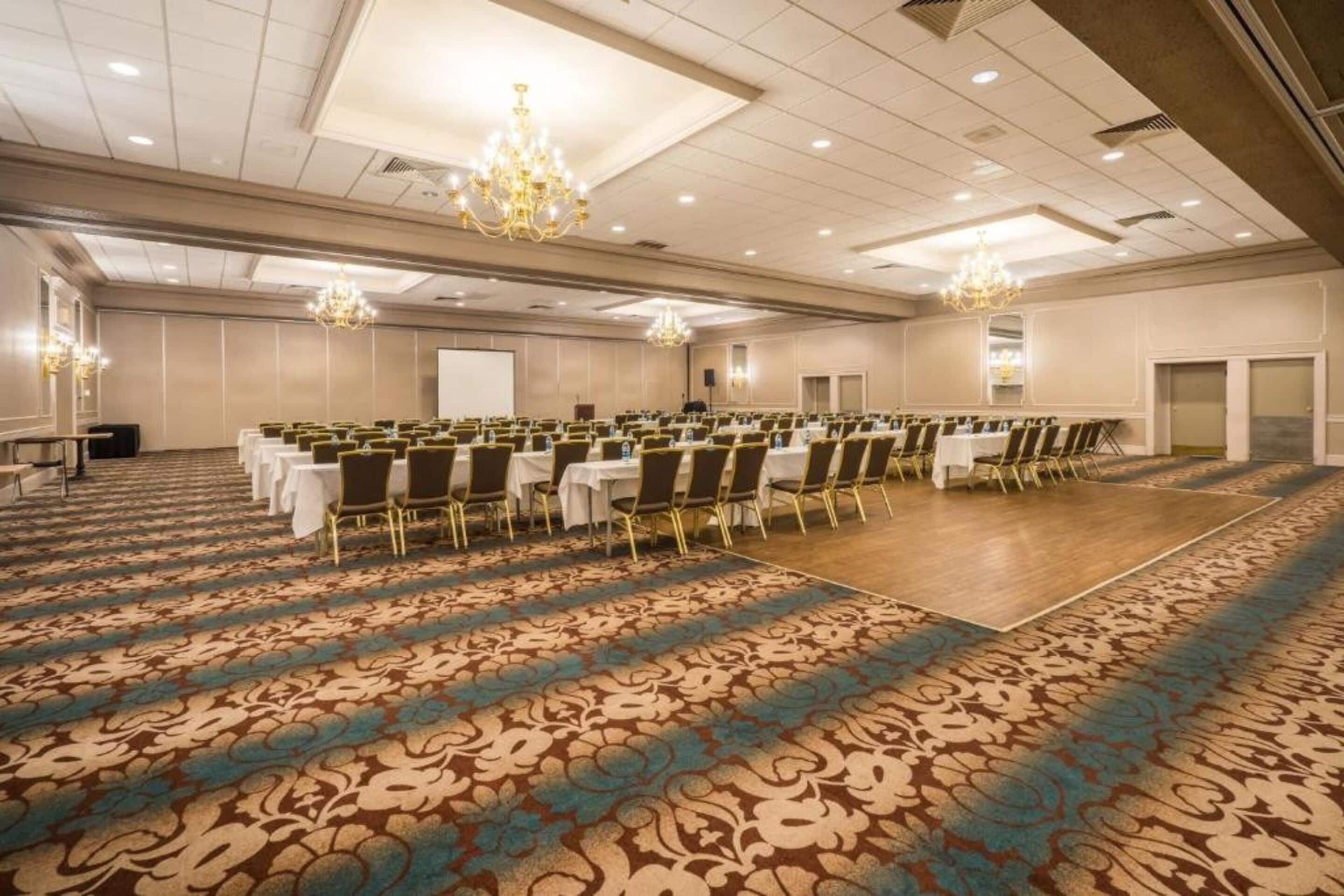 Une grande salle de conférence avec des rangées de tables et de chaises, une moquette à motifs, des lustres et un tableau blanc.