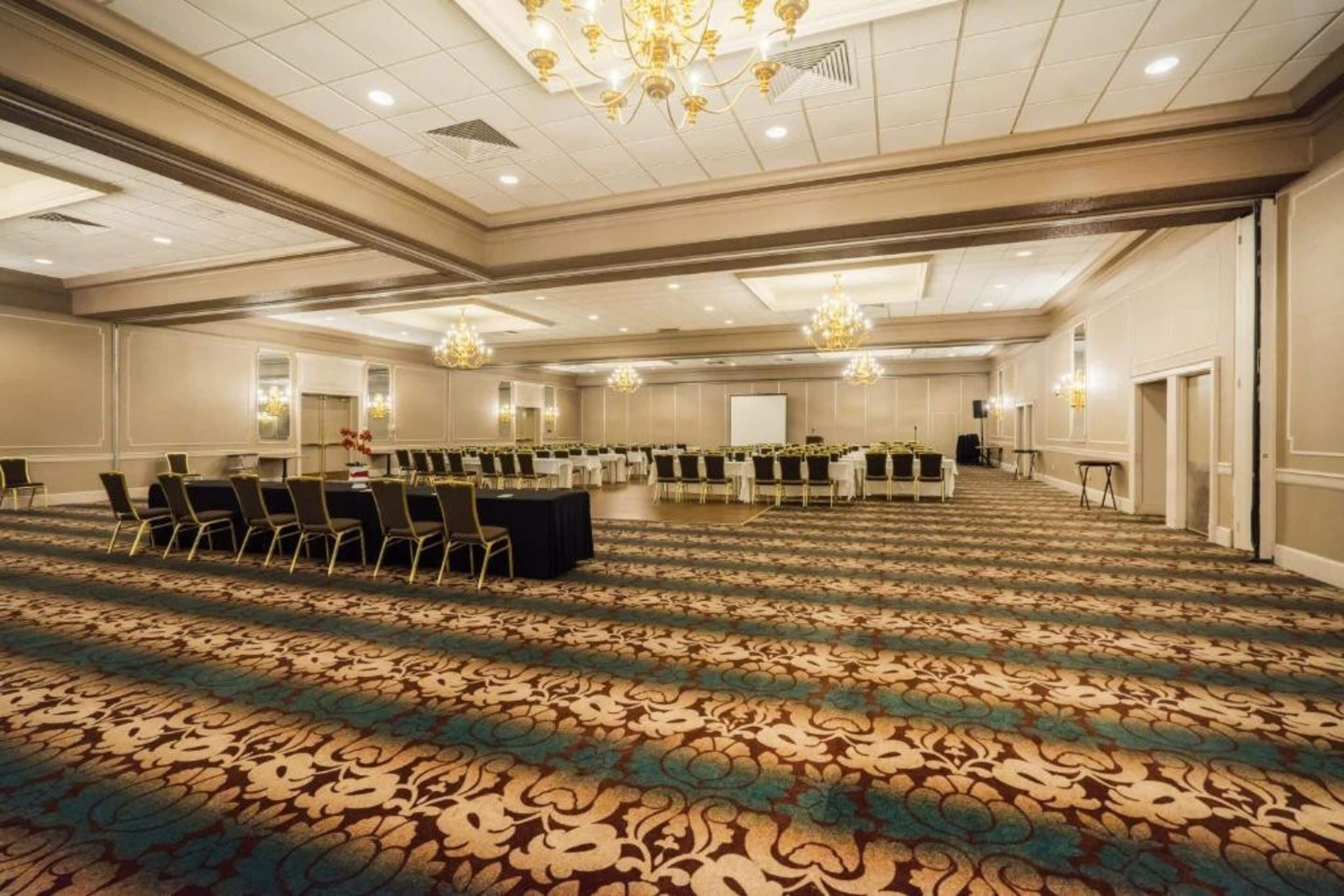 A large conference room with rows of tables and chairs, a patterned carpet, chandeliers, and a whiteboard.