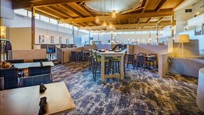 A spacious indoor area with high ceilings, exposed beams, and hanging pendant lights. There are multiple seating areas with tables and chairs, and a central table with a few chairs. The floor has a patterned carpet, and there are windows allowing natural light to enter. - enVision Hotel & Conference Center Mansfield-Foxboro (Mansfield)