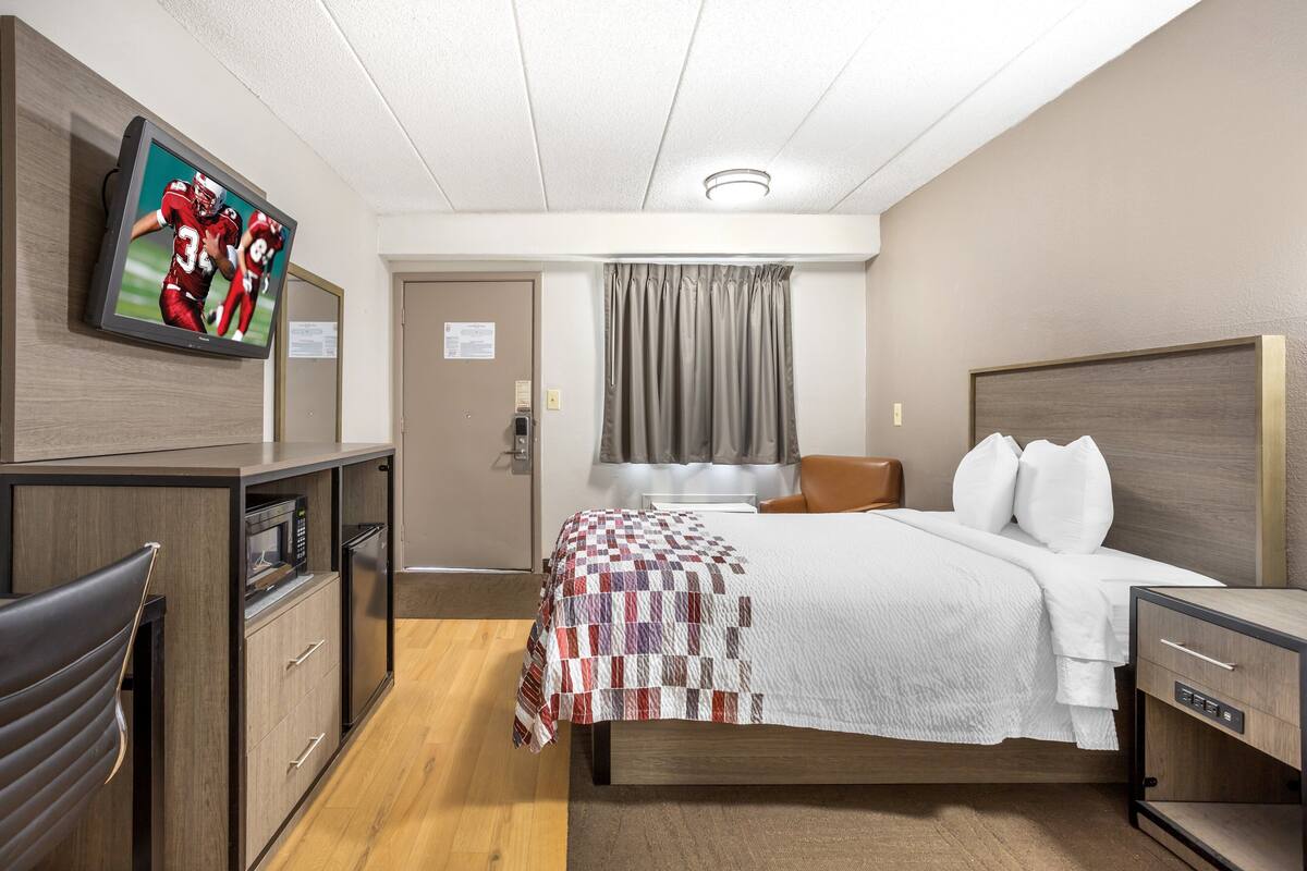 Habitación de hotel con cama, televisión montada en la pared, escritorio con microondas y una silla.