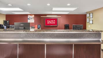 Zona de recepción de hotel con una pared roja, una computadora Lenovo, un dispensador de agua y un letrero que ofrece Wi-Fi gratuito en todas las habitaciones.