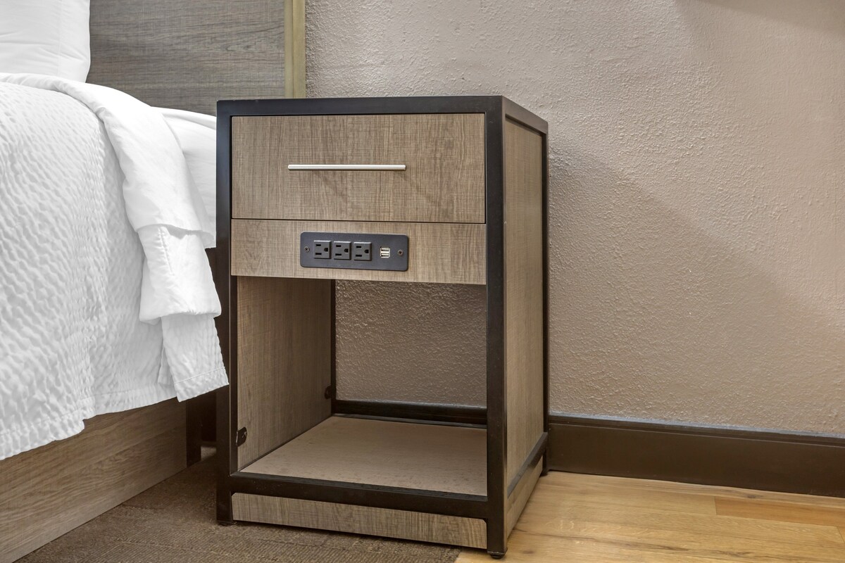A bedside table with a drawer and a power outlet, positioned next to a bed with a white textured bedspread.