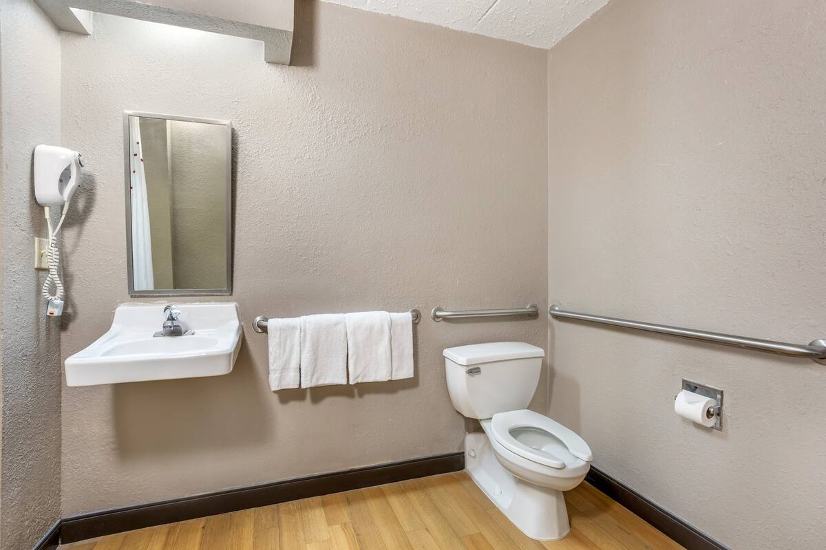 Un baño con lavamanos, espejo, toallero, inodoro y secamanos.