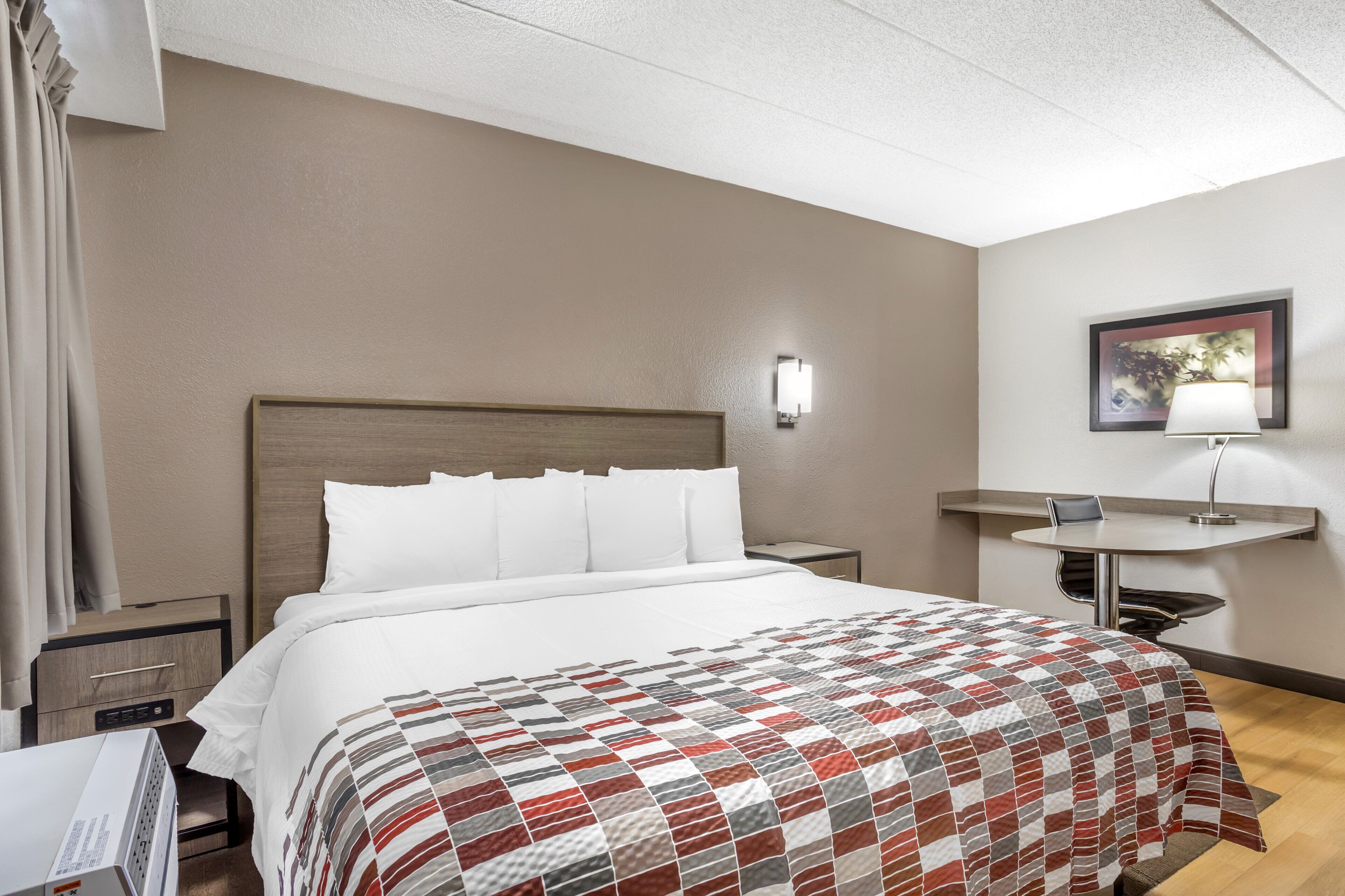 A hotel room with a bed, a desk, a chair, a nightstand, a lamp, and a picture on the wall.