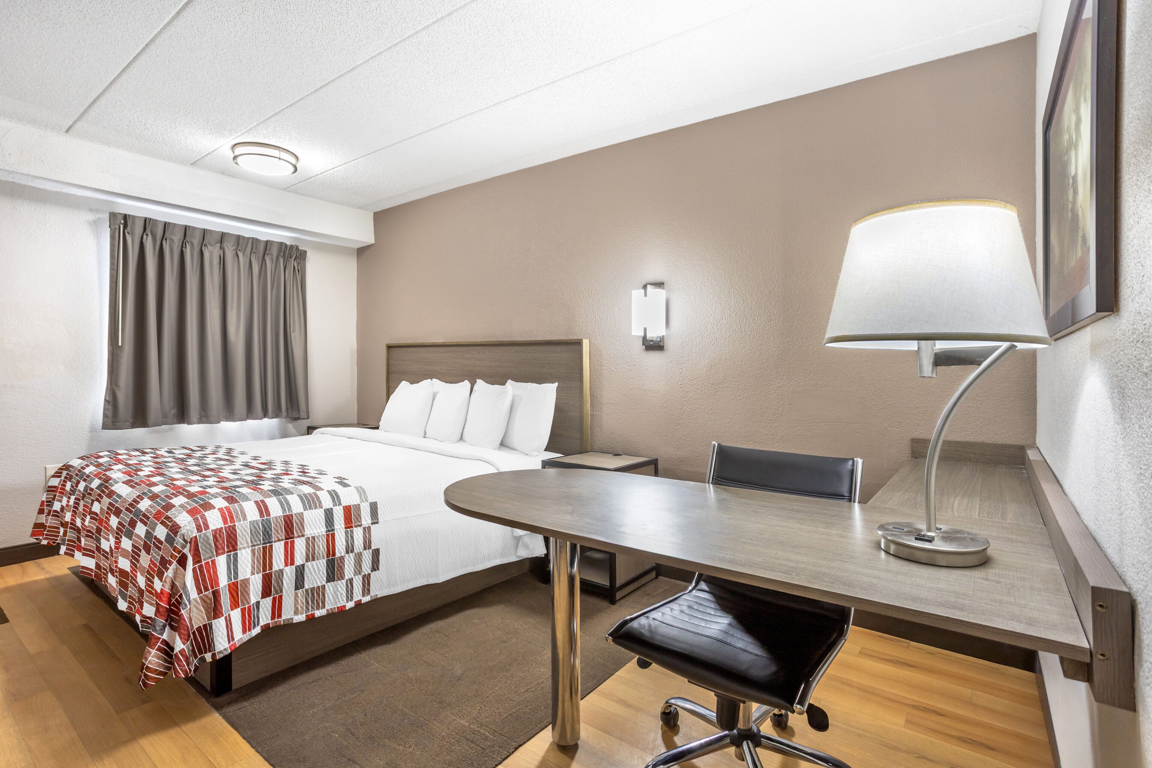 Habitación de hotel con cama, escritorio, silla, lámpara y un cuadro en la pared.