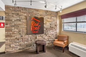 Ein Innenraum eines Red Roof Inn mit einer Steinwand, einem Schild, einem Ledersessel, einem kleinen Tisch und einem Fenster mit Jalousien.