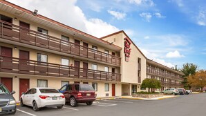 A multi-story hotel with a red and beige facade, balconies, and a parking lot with several cars. - Red Roof Inn Tinton Falls - Jersey Shore (Tinton Falls)