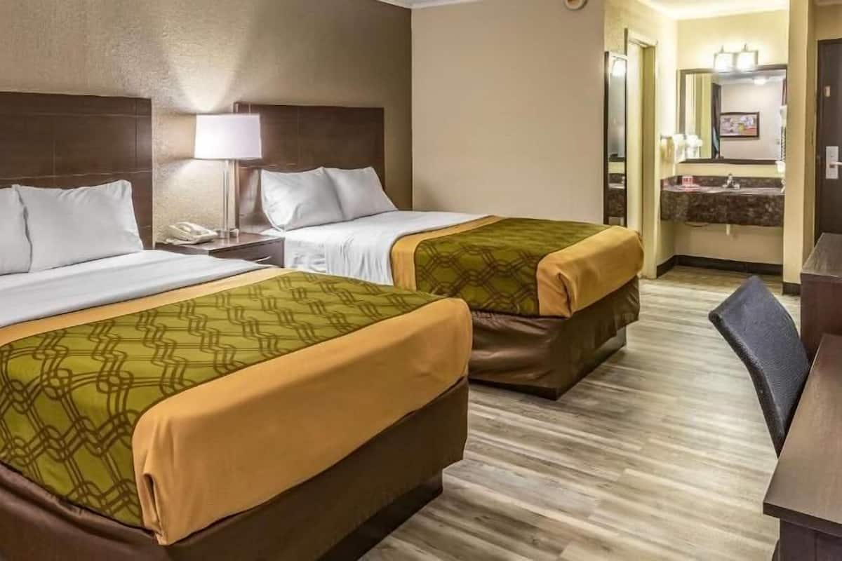 A hotel room with two beds, a nightstand, a lamp, a desk, and a bathroom visible through an open door.