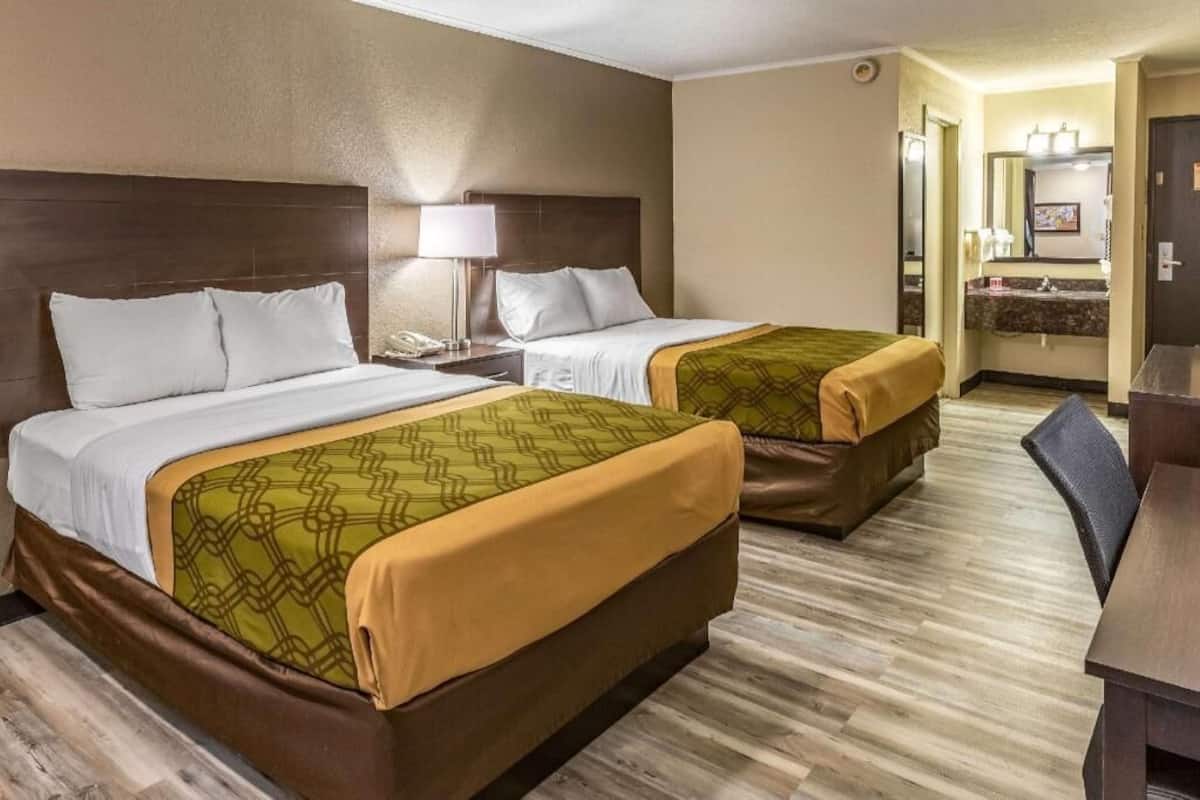 A hotel room with two beds, a desk, a chair, a bathroom with a mirror, and a sink.