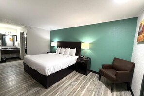 A hotel room with a bed, bedside table, lamp, chair, and a bathroom with a mirror and sink. - Baymont Inn & Suites by Wyndham Newton IA (Newton)
