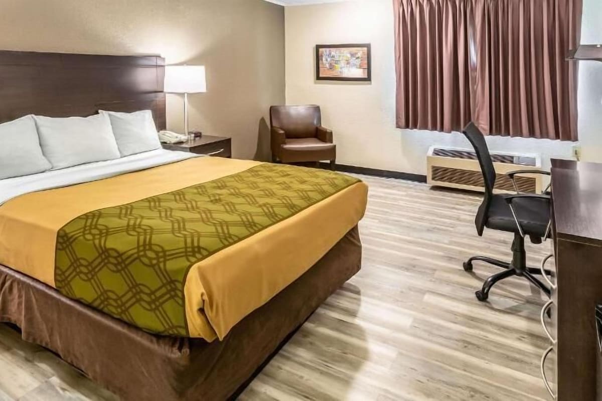 A hotel room with a bed, a desk, a chair, a lamp, and a painting on the wall.
