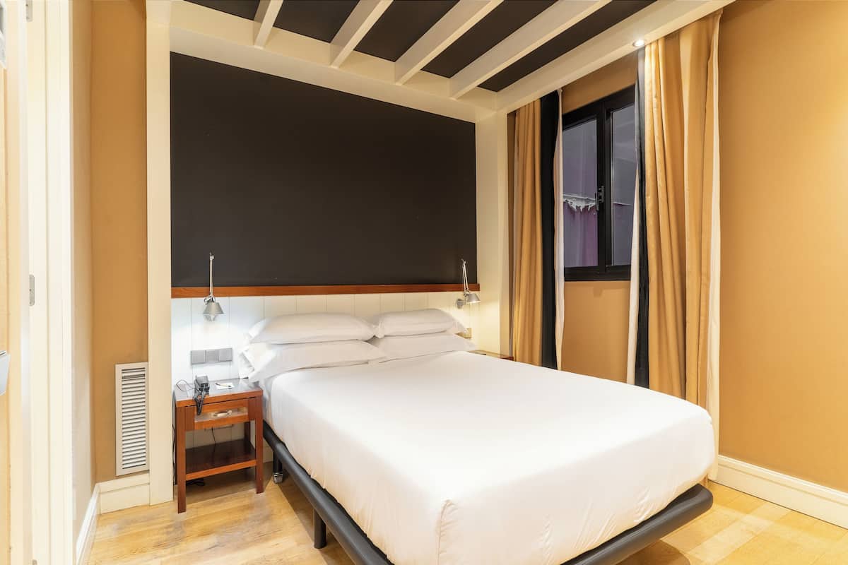 Una cama bien tendida con sábanas blancas, una mesita de noche con lámpara y una ventana con cortinas.