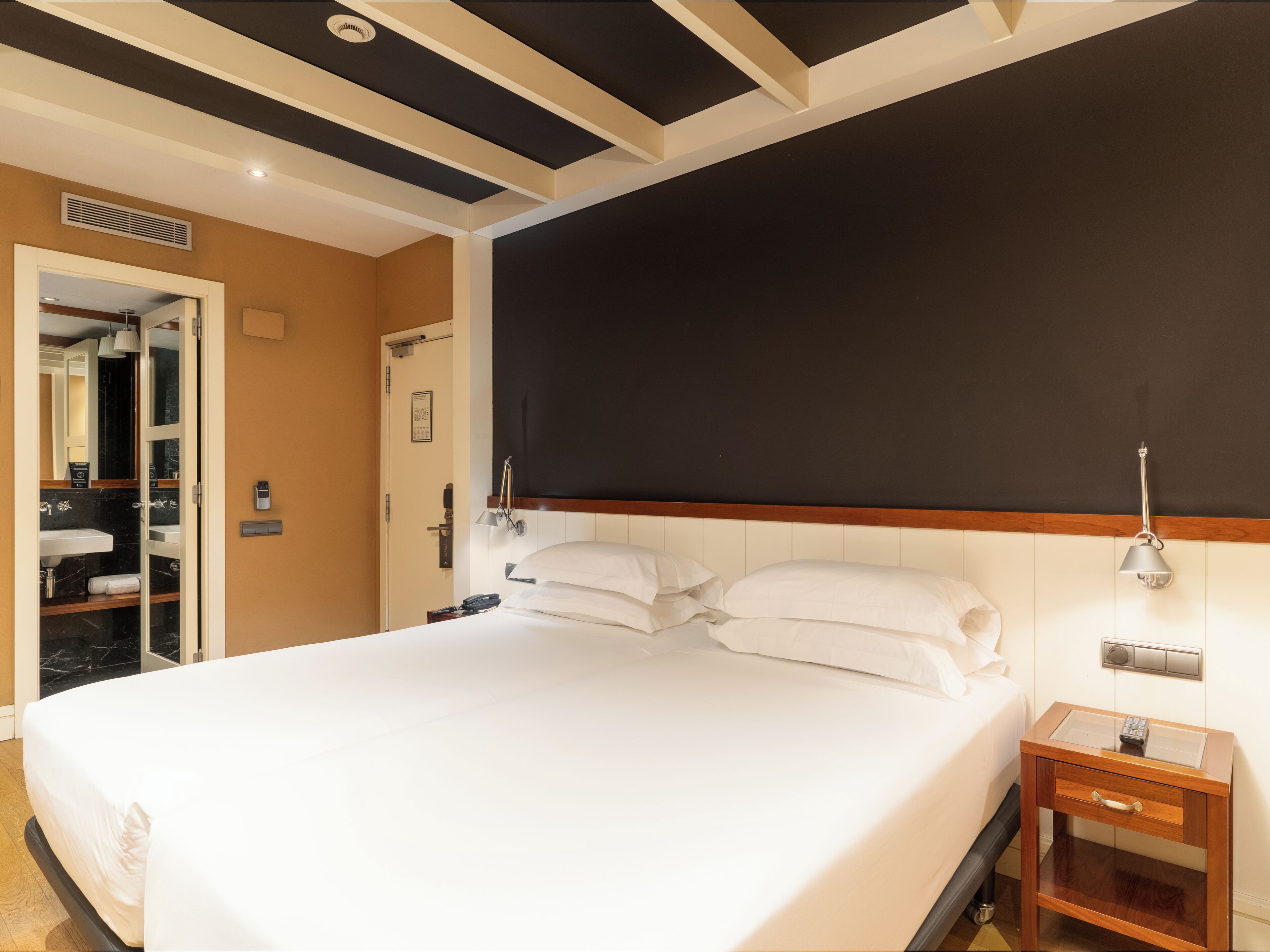 Una habitación de hotel con una cama grande, una mesita de noche con lámpara, una mesita auxiliar de madera y un baño visible a través de una puerta abierta.