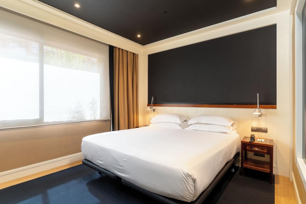 Habitación de hotel con una cama grande, una mesita de noche, una lámpara de pared y una ventana con cortinas.