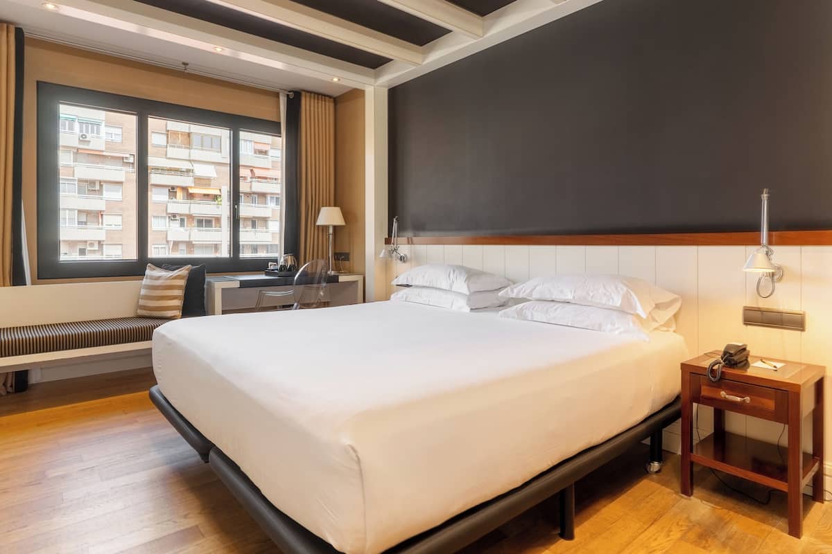 Una habitación de hotel con una cama grande, un escritorio con una silla, una mesita de noche con lámpara y vistas a la ciudad desde la ventana.