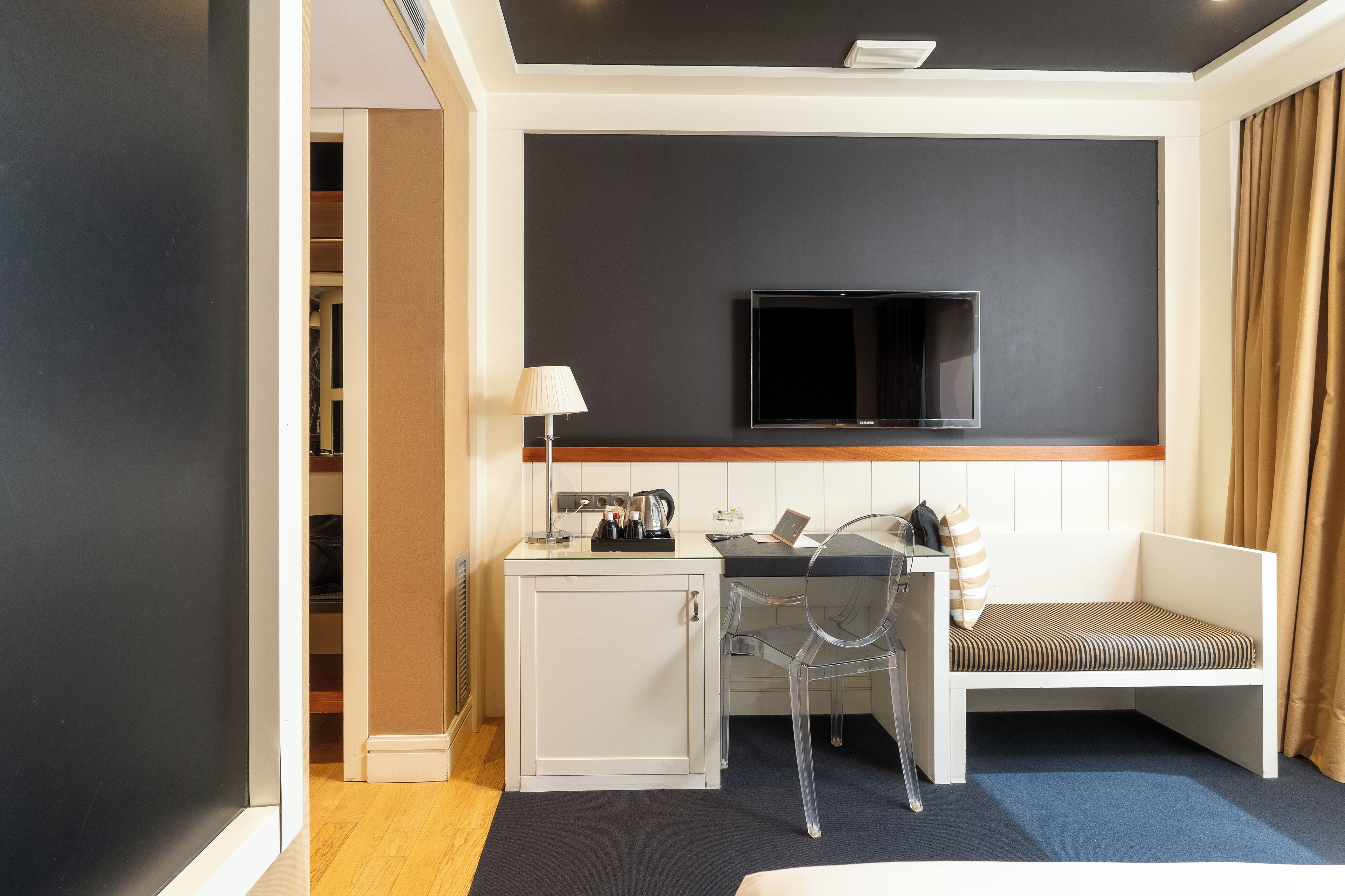 A compact hotel room with a kitchenette, a TV mounted on the wall, a seating area with a bench, and a desk with a chair.