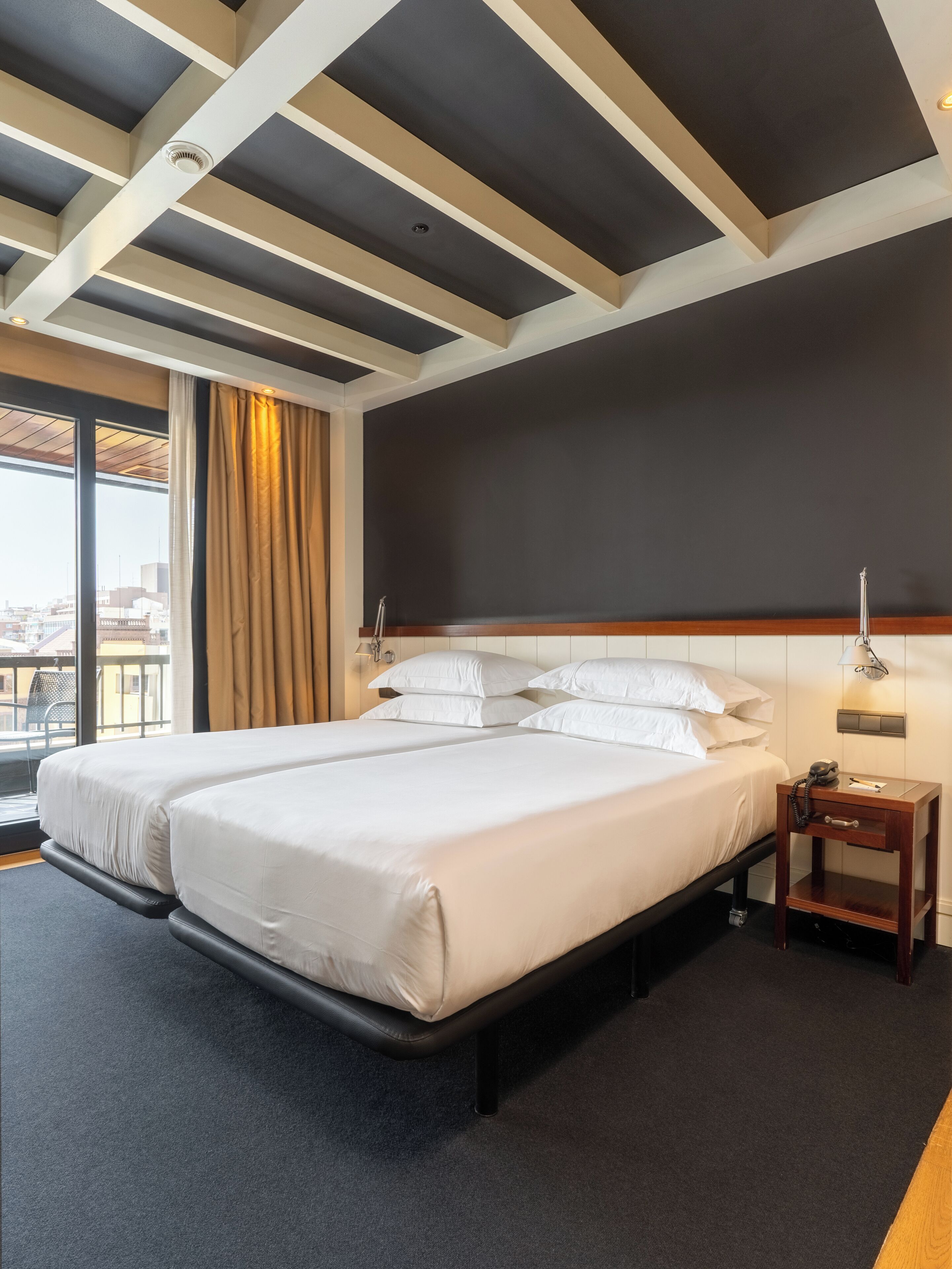 Habitación de hotel con una cama grande, una mesita de noche con lámpara y vistas a la ciudad a través de la ventana.