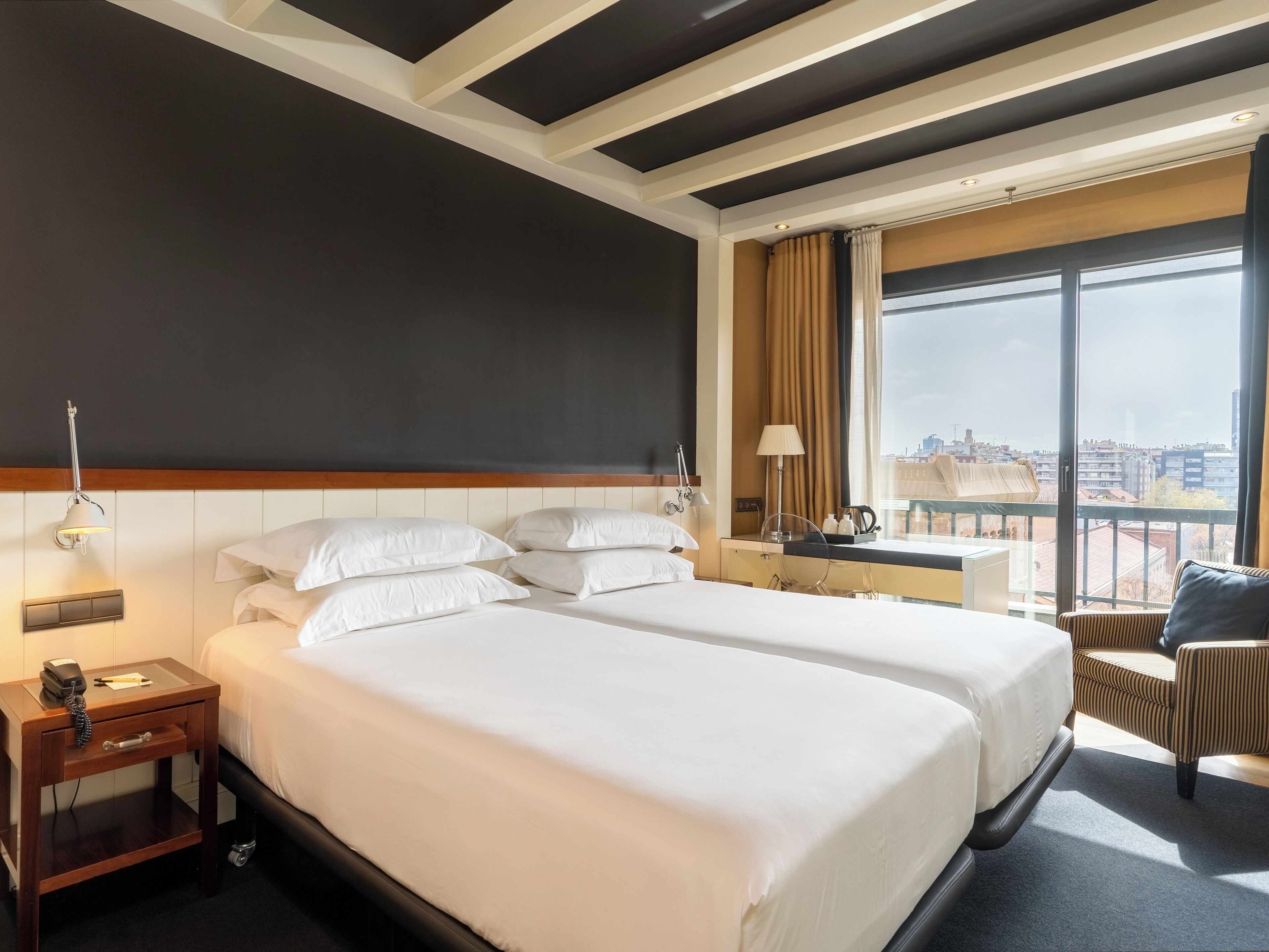 A hotel room with two beds, a desk, a chair, a large window with a city view, and a modern ceiling design.