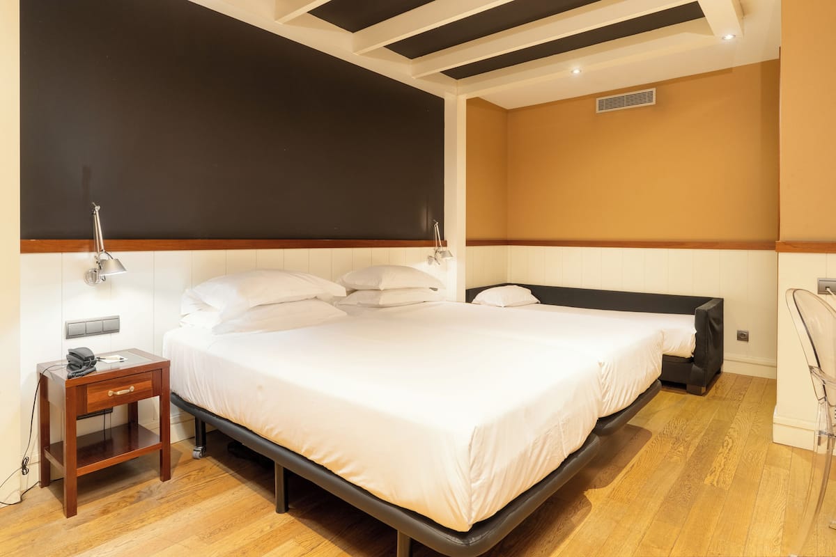 Una cama doble con cabecera de madera, una mesita de noche con lámpara, y otro mueble auxiliar con teléfono y cargador.
