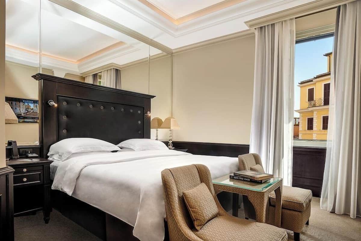 A hotel room with a large bed, a small table, a chair, a nightstand, and a window with curtains.