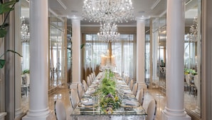 Une longue table à manger, dressée pour un événement formel, flanquée de hautes colonnes, et surmontée d’un grand lustre.