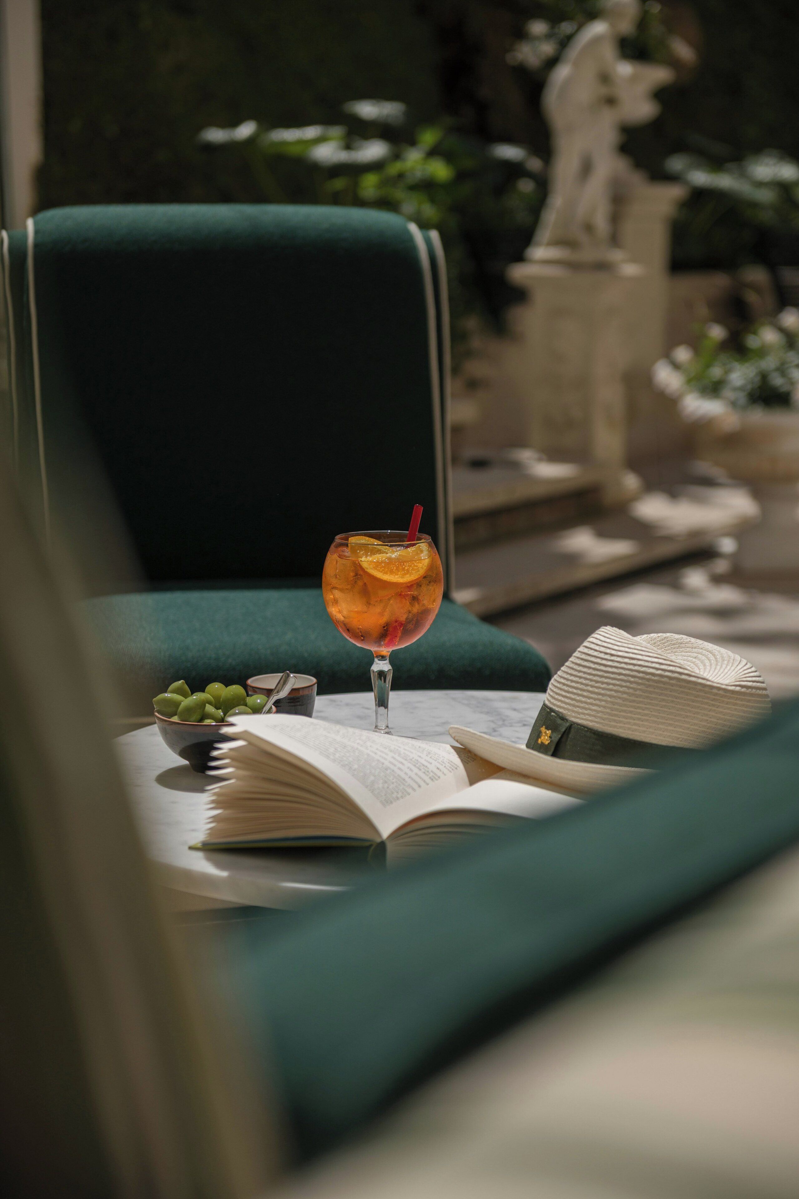 A glass of drink with an orange slice on a table with an open book and a bowl of grapes.