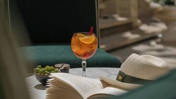 A glass of drink with an orange slice on a table with an open book and a bowl of grapes.