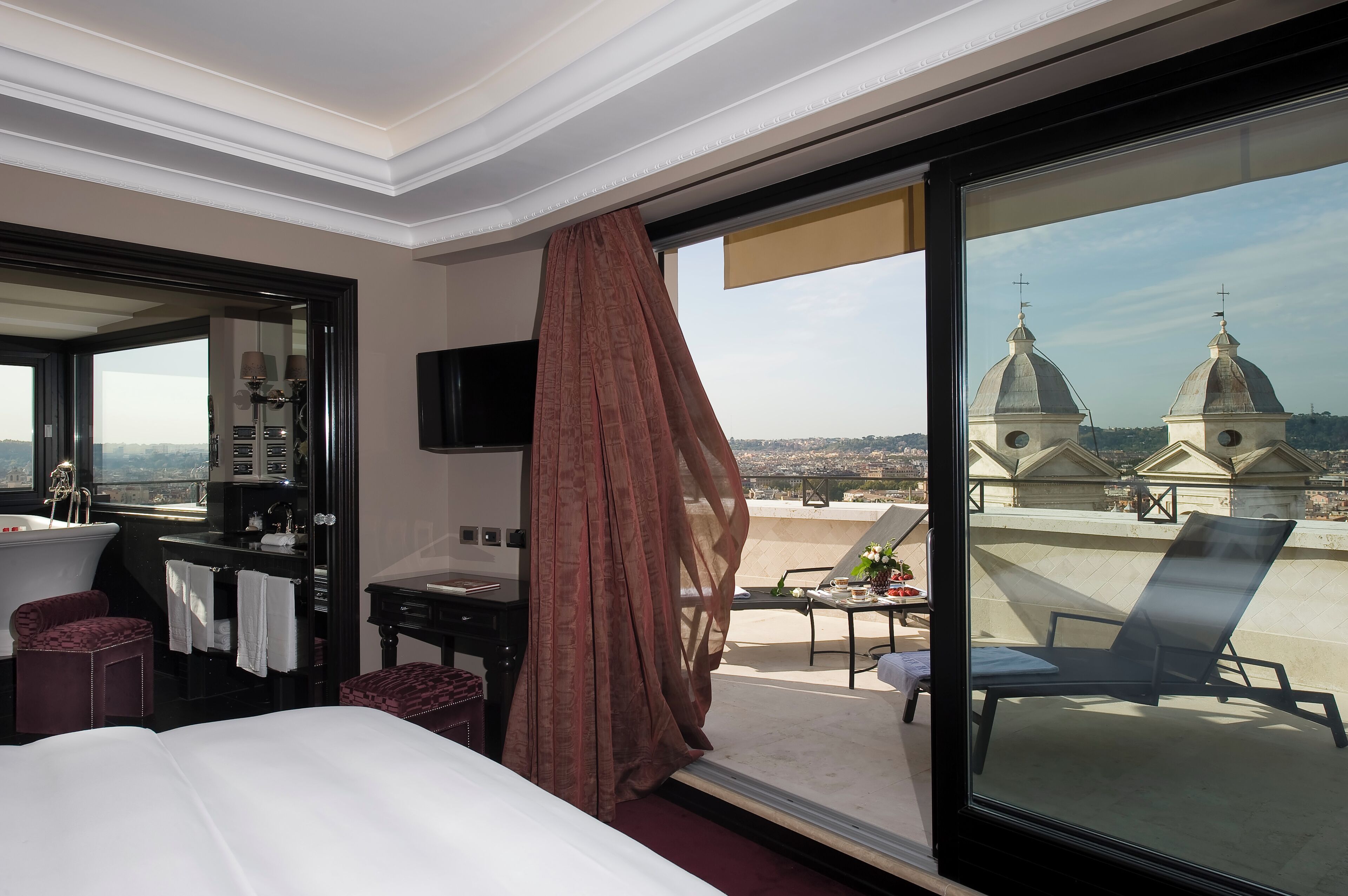 A hotel room with a large bed, a desk, a chair, and a balcony with a view of domed buildings.