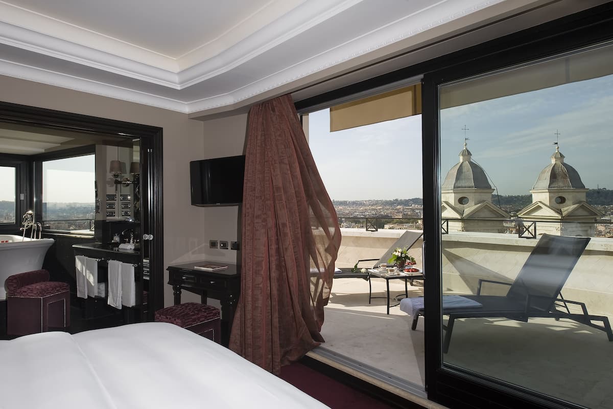 A hotel room with a large bed, a desk, a chair, and a balcony with a view of domed buildings.