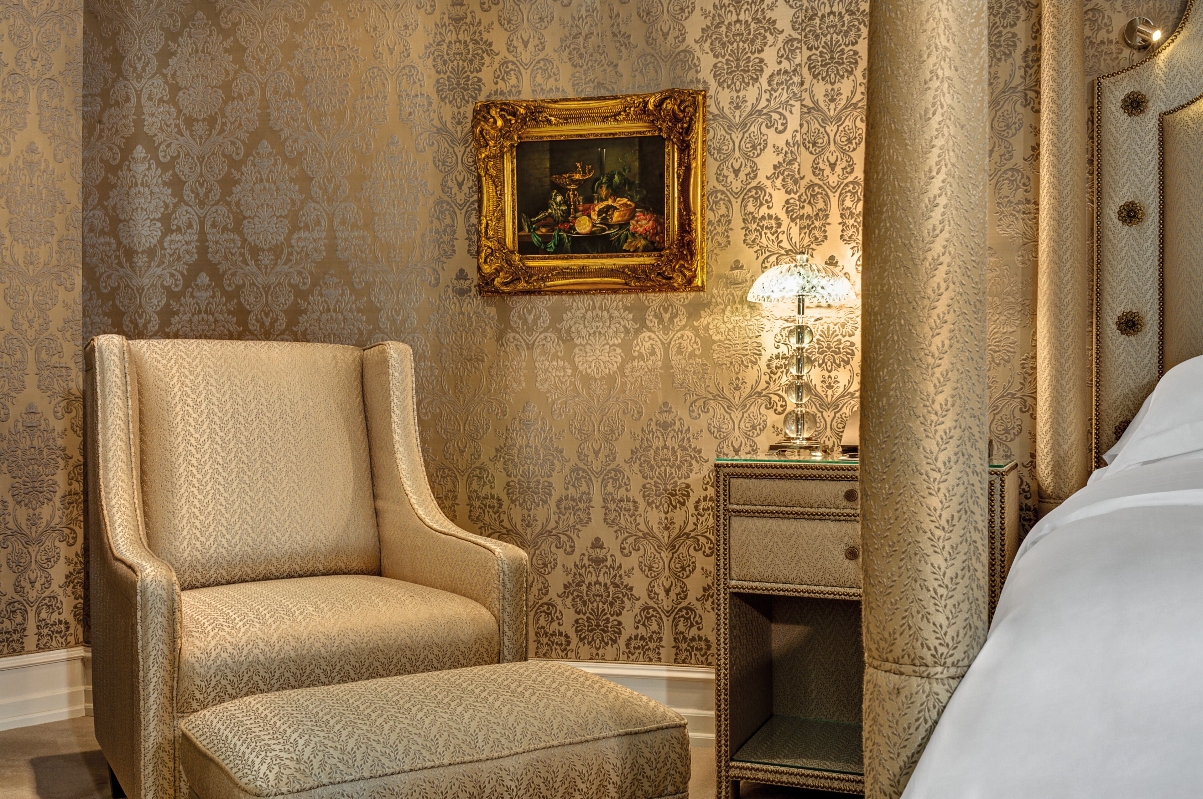A hotel room with a patterned wallpaper, a framed painting on the wall, a bedside table with a lamp, and a large bed.